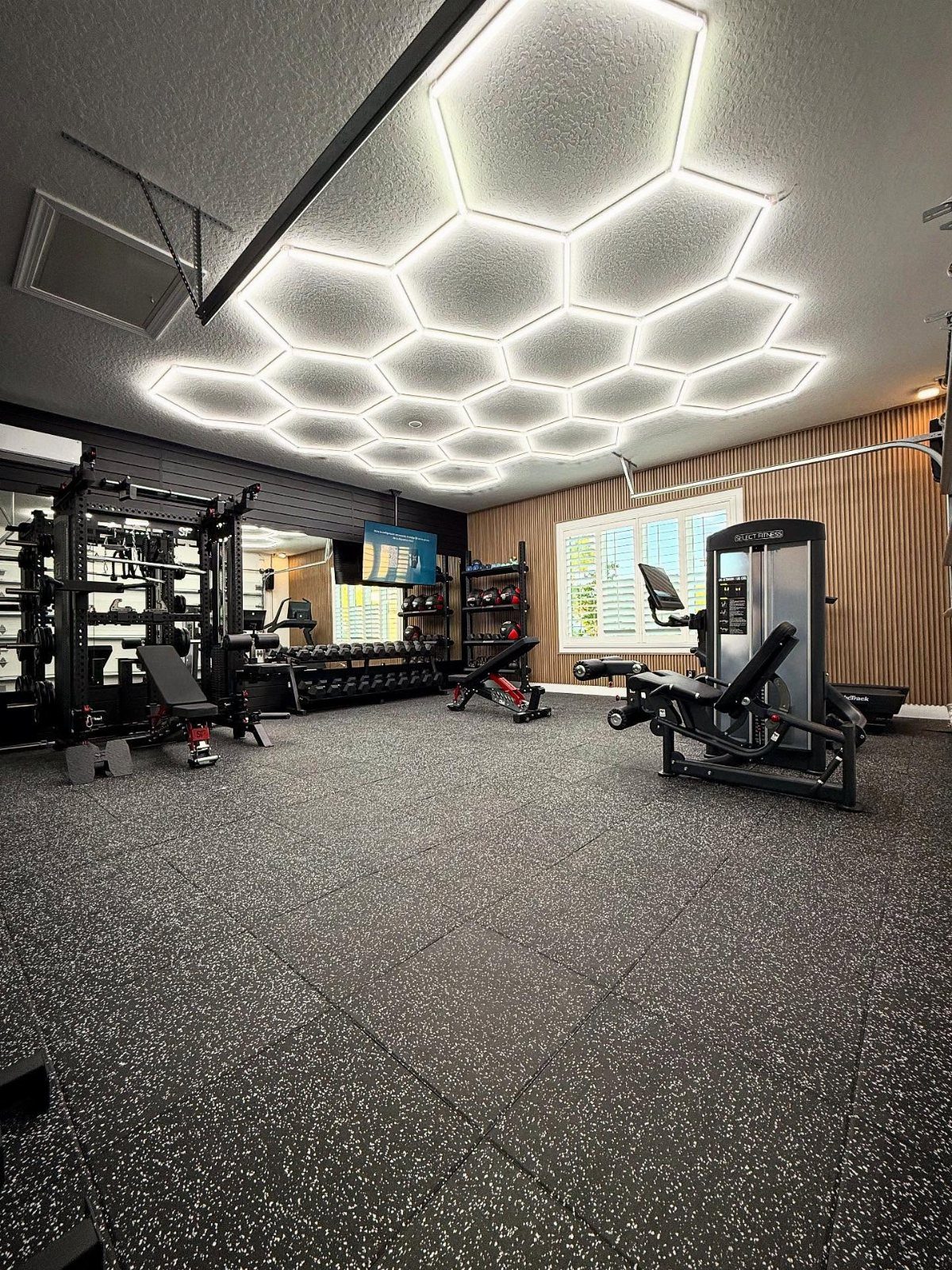 Black gym equipment and weight racks arranged in a room with hexagonal ceiling lights.