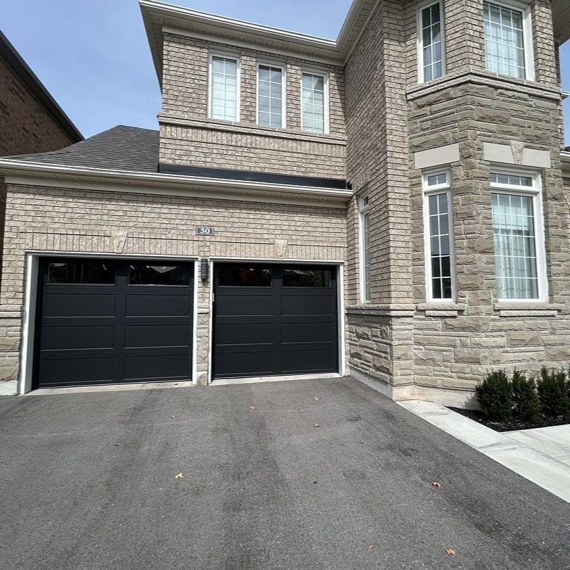 A two-car garage with black doors on a stone and brick house with an asphalt driveway.