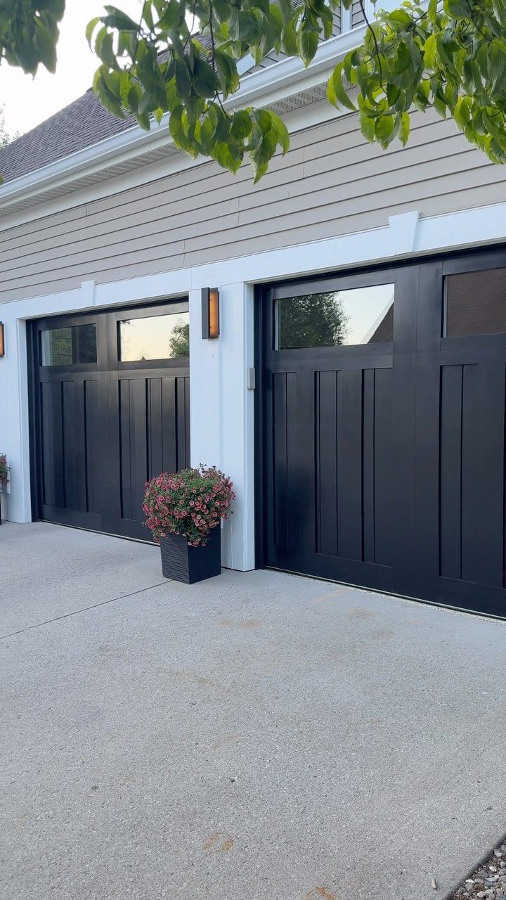 Two black modern garage doors with vertical panels and windows on a light grey house.