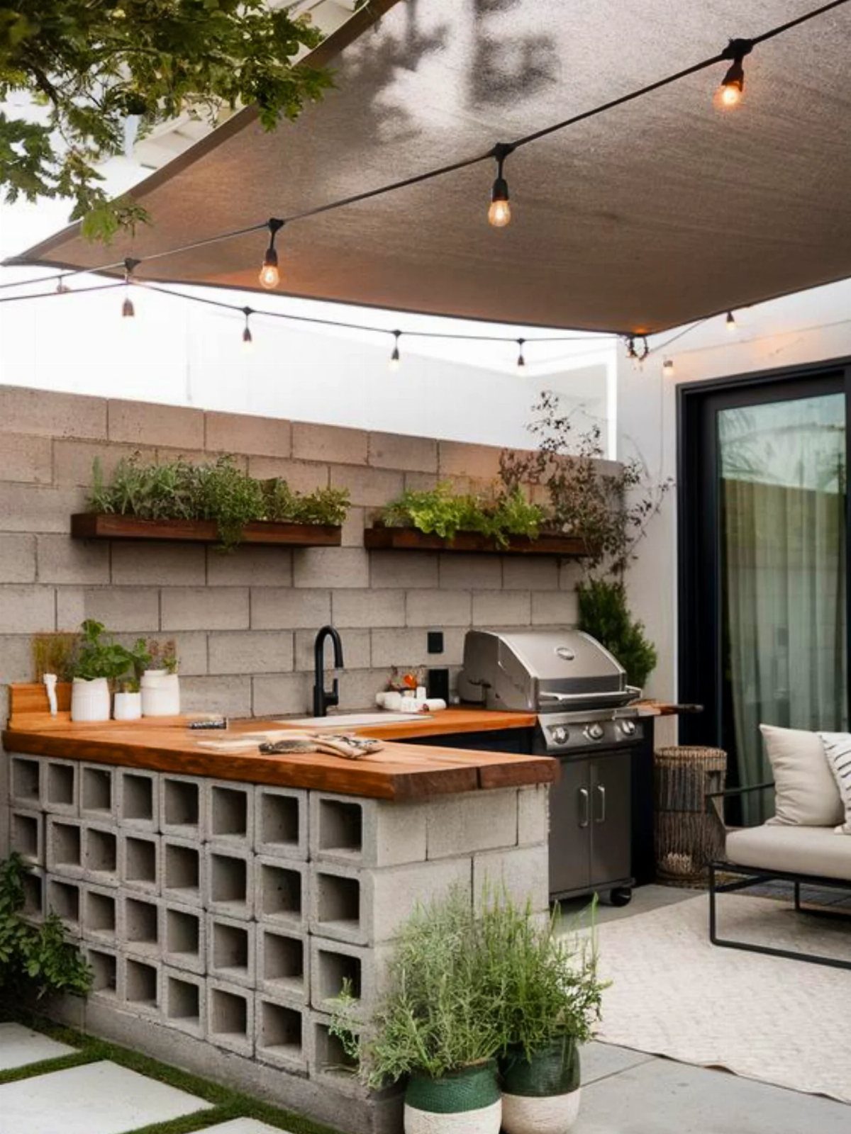 Concrete block BBQ island with wooden countertops, stainless steel grill, and herb planters in a backyard.
