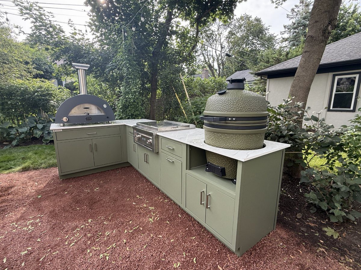 Olive green outdoor kitchen cabinetry with a pizza oven, grill, and ceramic smoker on white countertops.