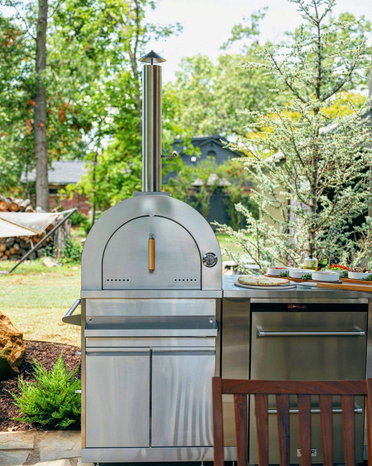 Stainless steel outdoor kitchen featuring a dome pizza oven and built-in grill in a backyard.