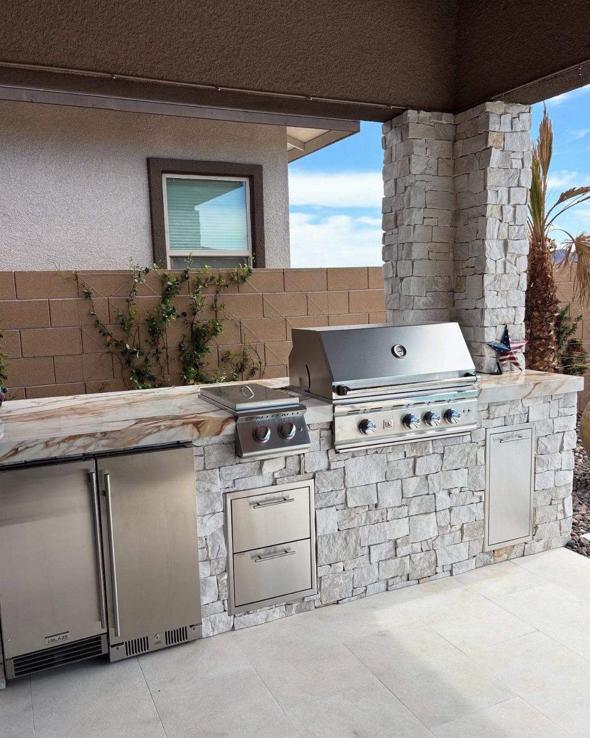 Stainless steel outdoor kitchen island with stone cladding and built-in appliances under a covered patio.