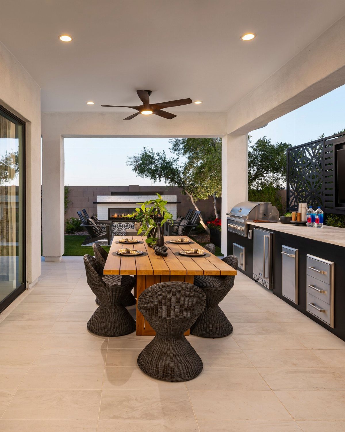 A wooden dining table with dark wicker chairs set on a tiled patio under a covered structure.