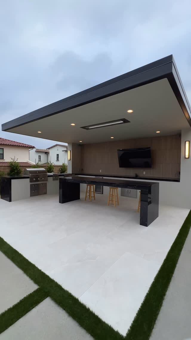 Modern outdoor kitchen with a built-in grill, long black counter, and bar stools under a roof.