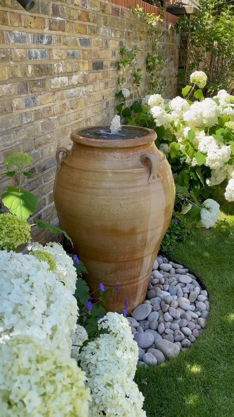 Large ceramic water urn fountain surrounded by white hydrangeas and river stones against a brick wall.