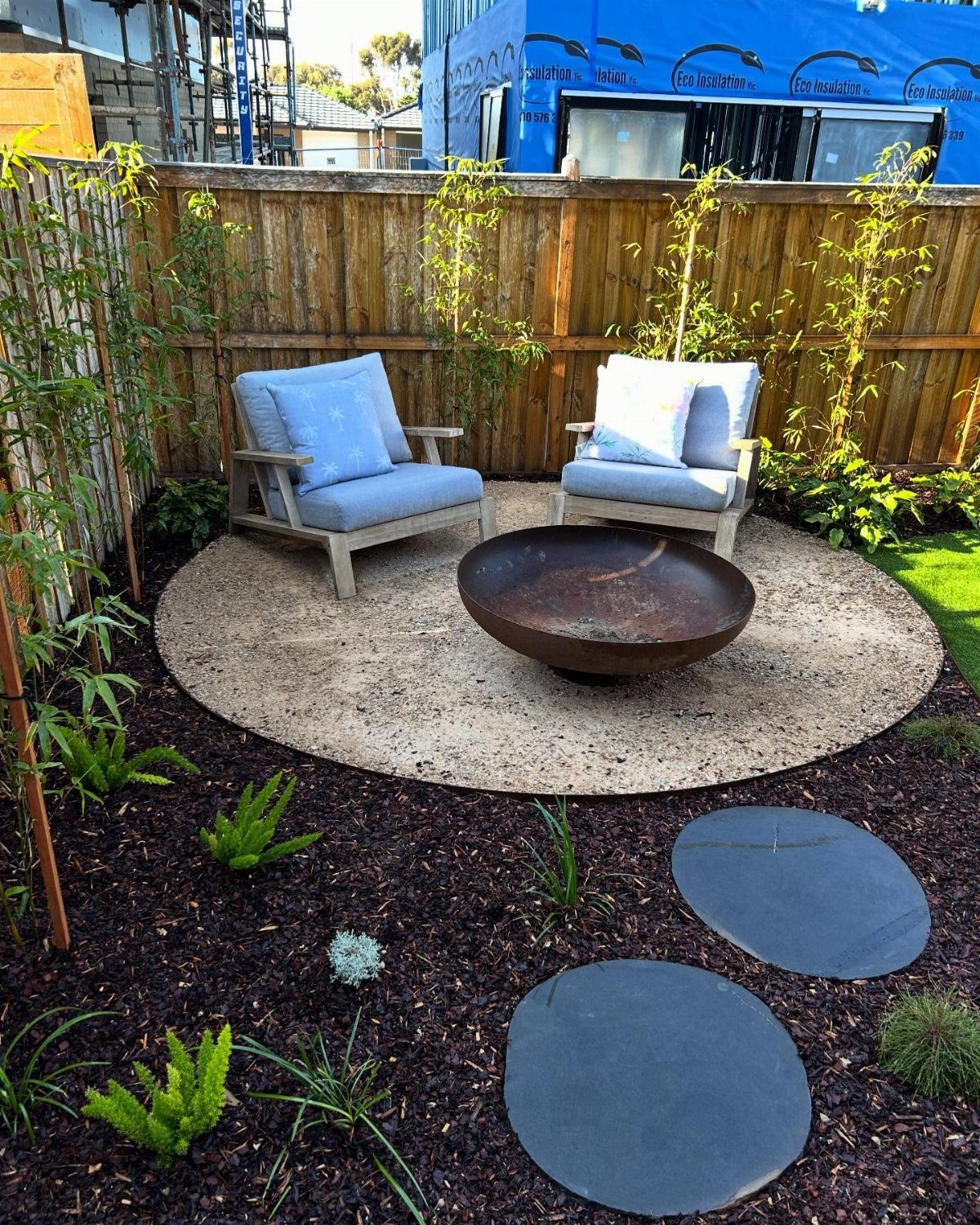 A metal fire bowl on a gravel patio surrounded by two blue cushioned chairs and garden plants.