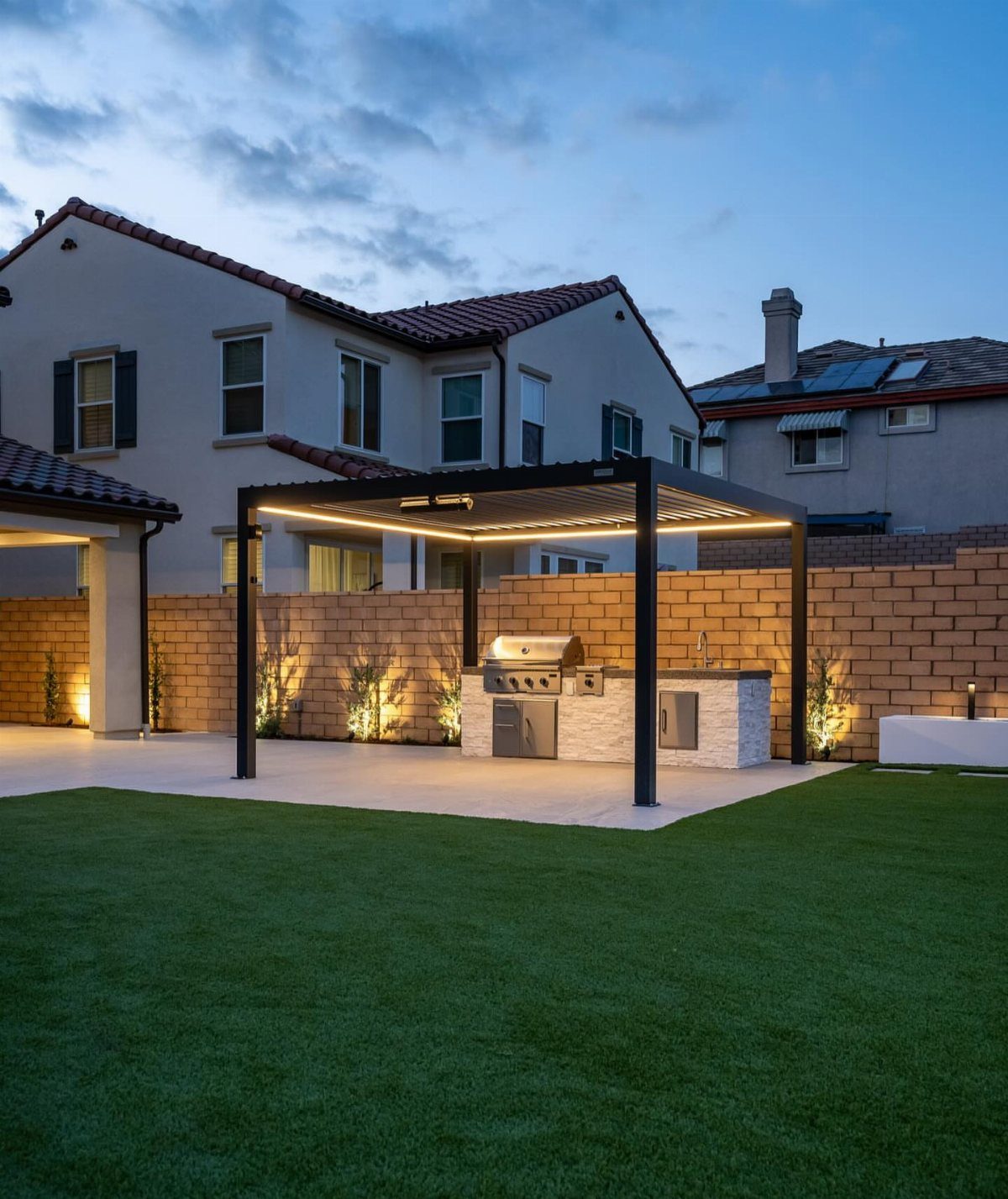 Modern outdoor kitchen with a black pergola and stainless steel grill on a paved patio.