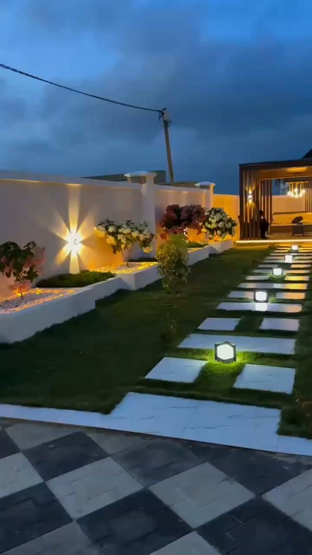 Square glowing lanterns illuminate a modern stone pathway through a manicured green lawn at dusk.