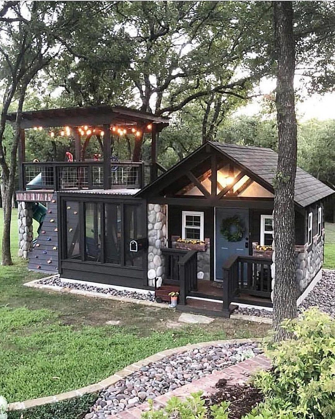 A dark wooden playhouse with stone walls, a porch, and a lit upper deck in a yard.
