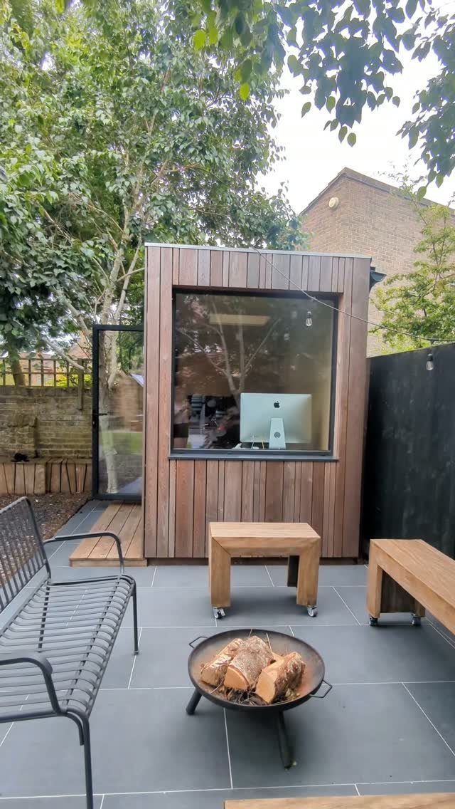 A wooden garden studio with a large window containing a computer on a patio.