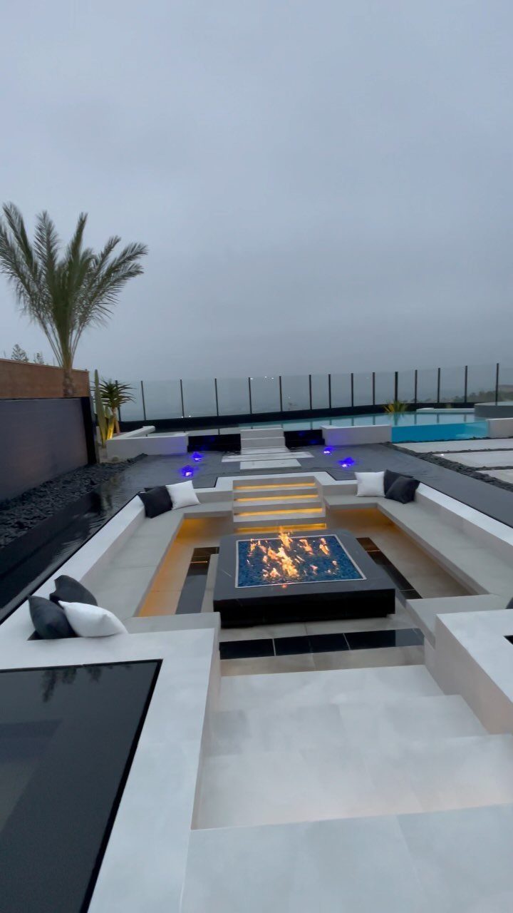 Modern outdoor lounge area with a square black fire pit and built-in stone seating.