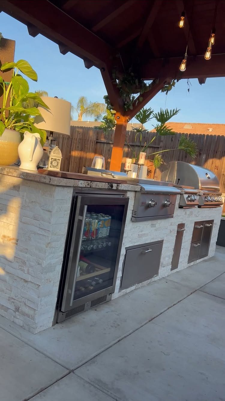 A built-in outdoor kitchen with stone tiling, a stainless steel grill, and a beverage fridge under a pergola.