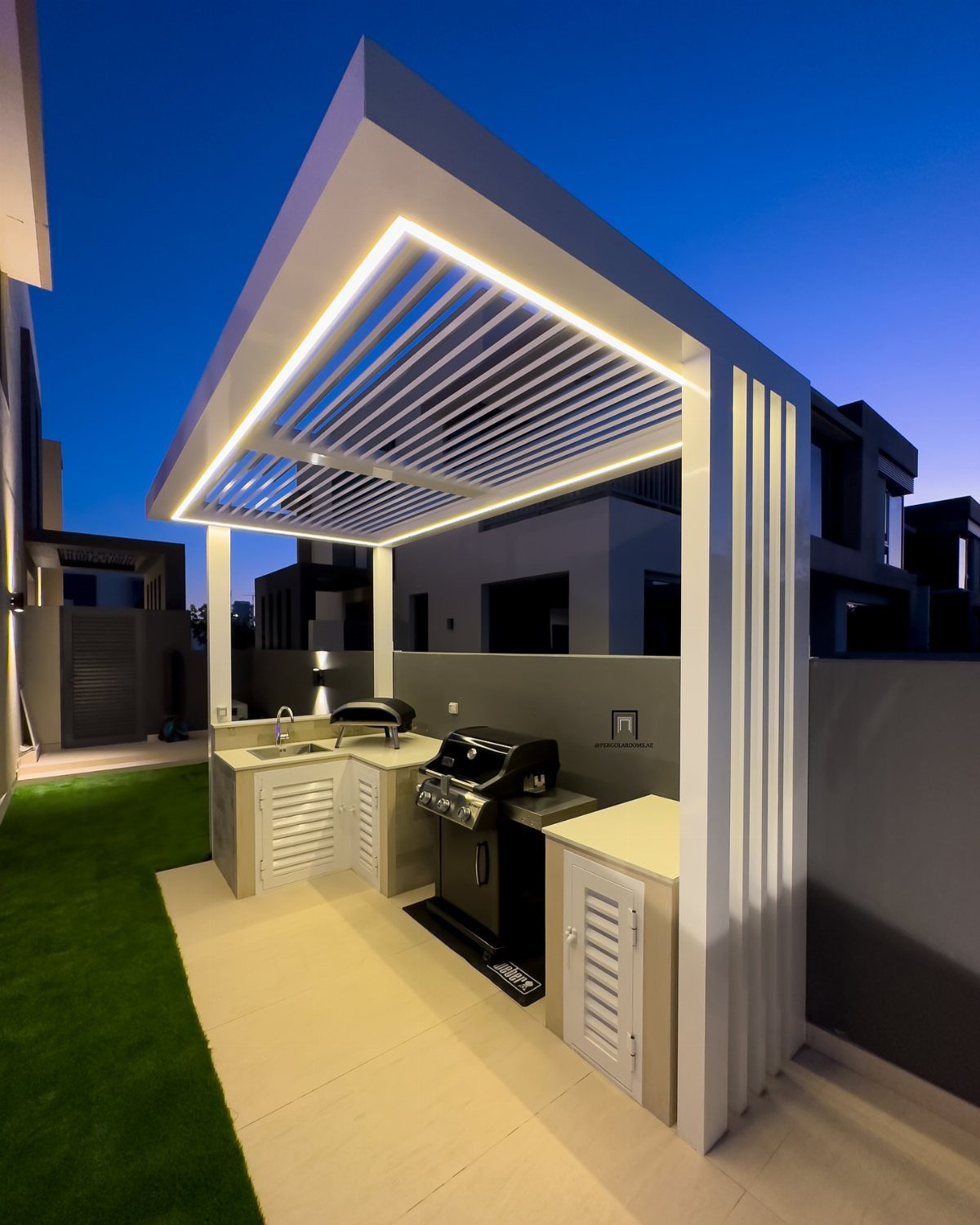 Modern outdoor kitchen with a built-in grill under a white illuminated pergola at dusk.