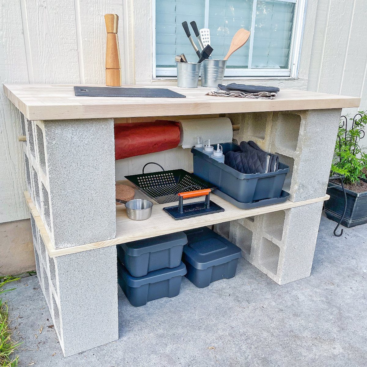 A DIY outdoor prep table constructed from cinder blocks and light wood planks holding kitchen supplies.