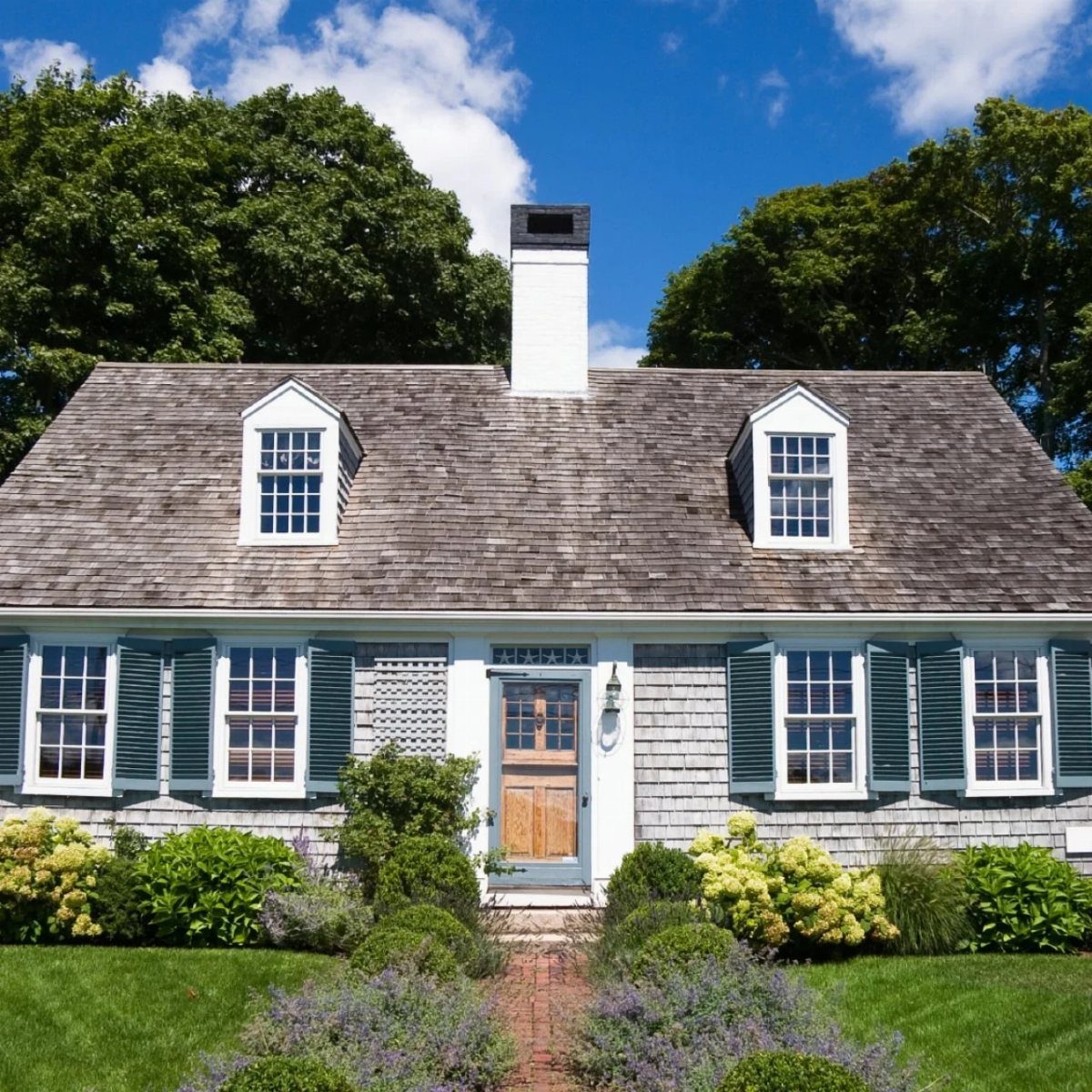 Grey shingle Cape Cod house featuring white trim, green shutters, and multiple double-hung windows.