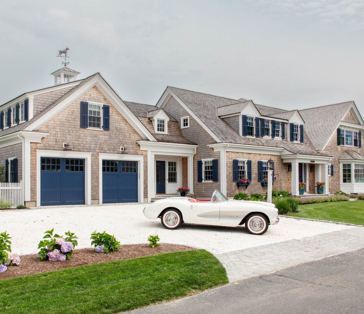 Grey shingle house with muted blue shutters and garage doors featuring a white vintage car.
