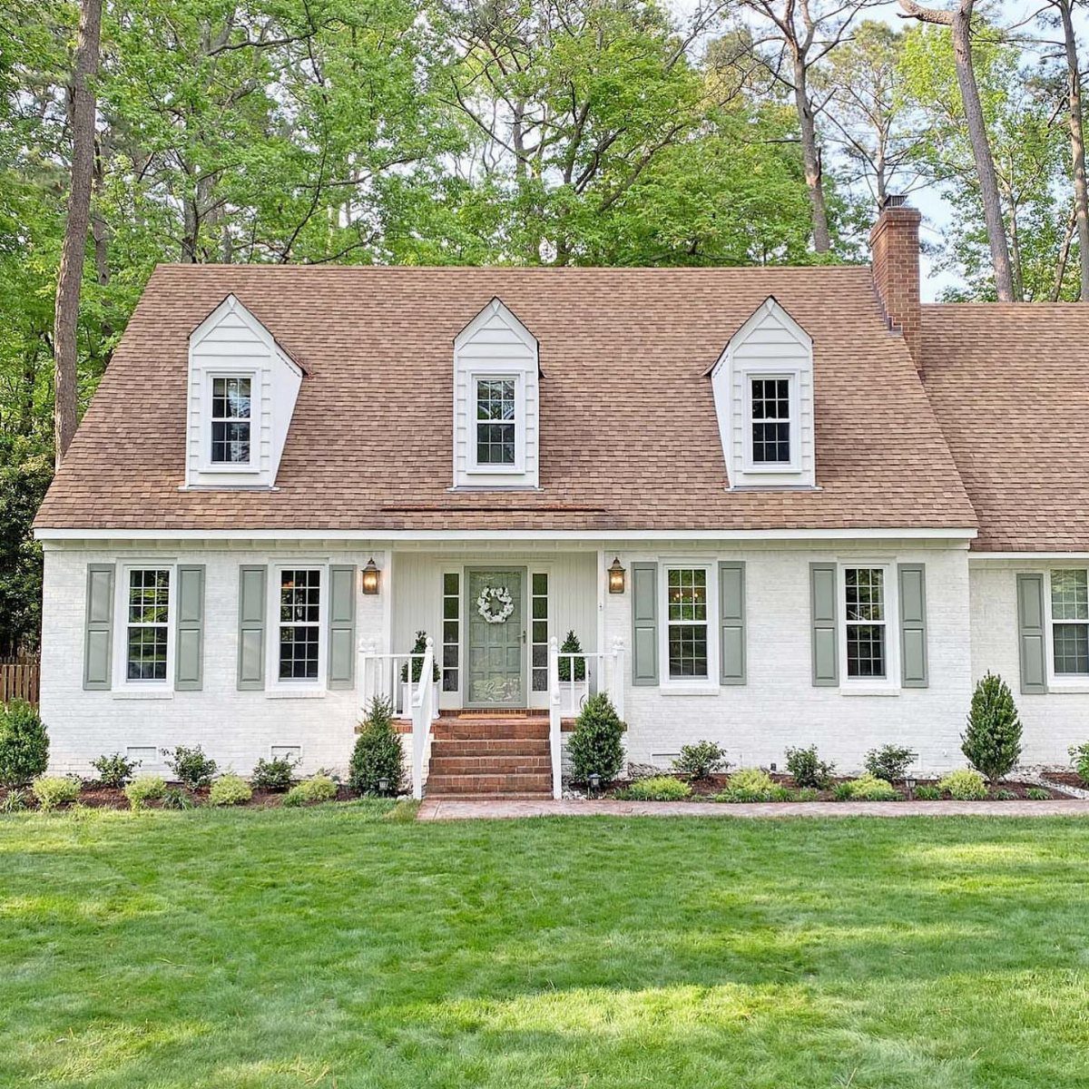White brick Cape Cod house featuring light green shutters and three dormer windows under a brown roof.