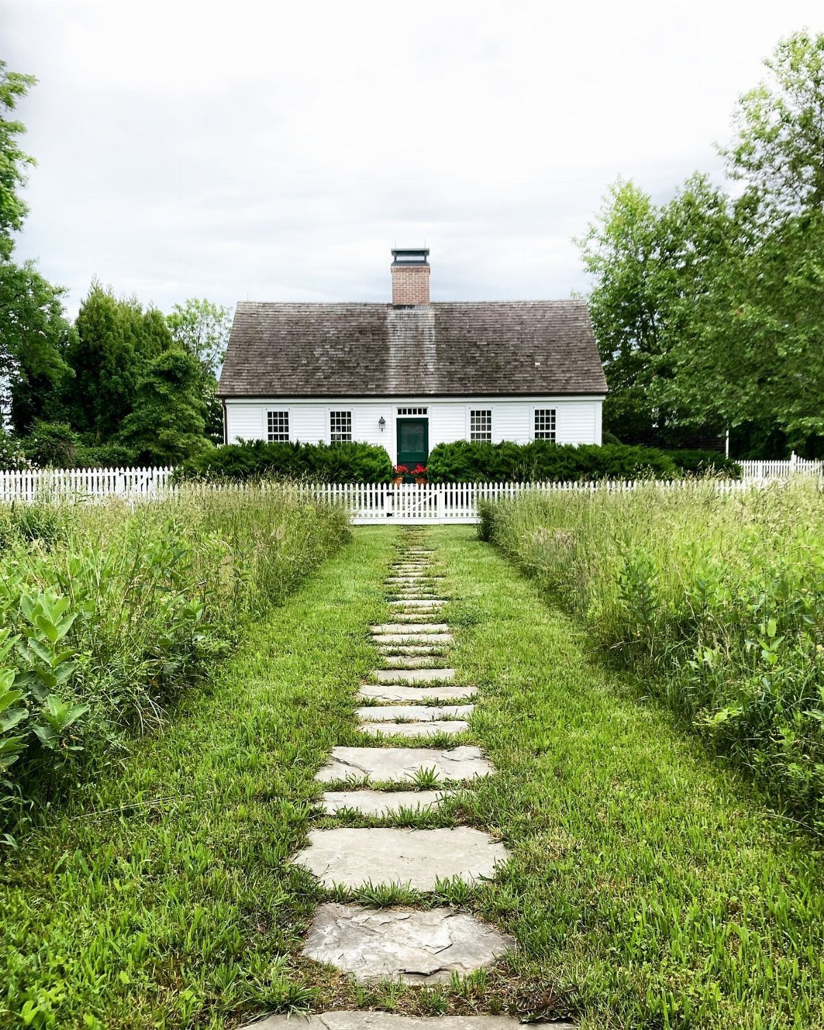 A white Cape Cod house with a green door sits behind a white picket fence and stone path.