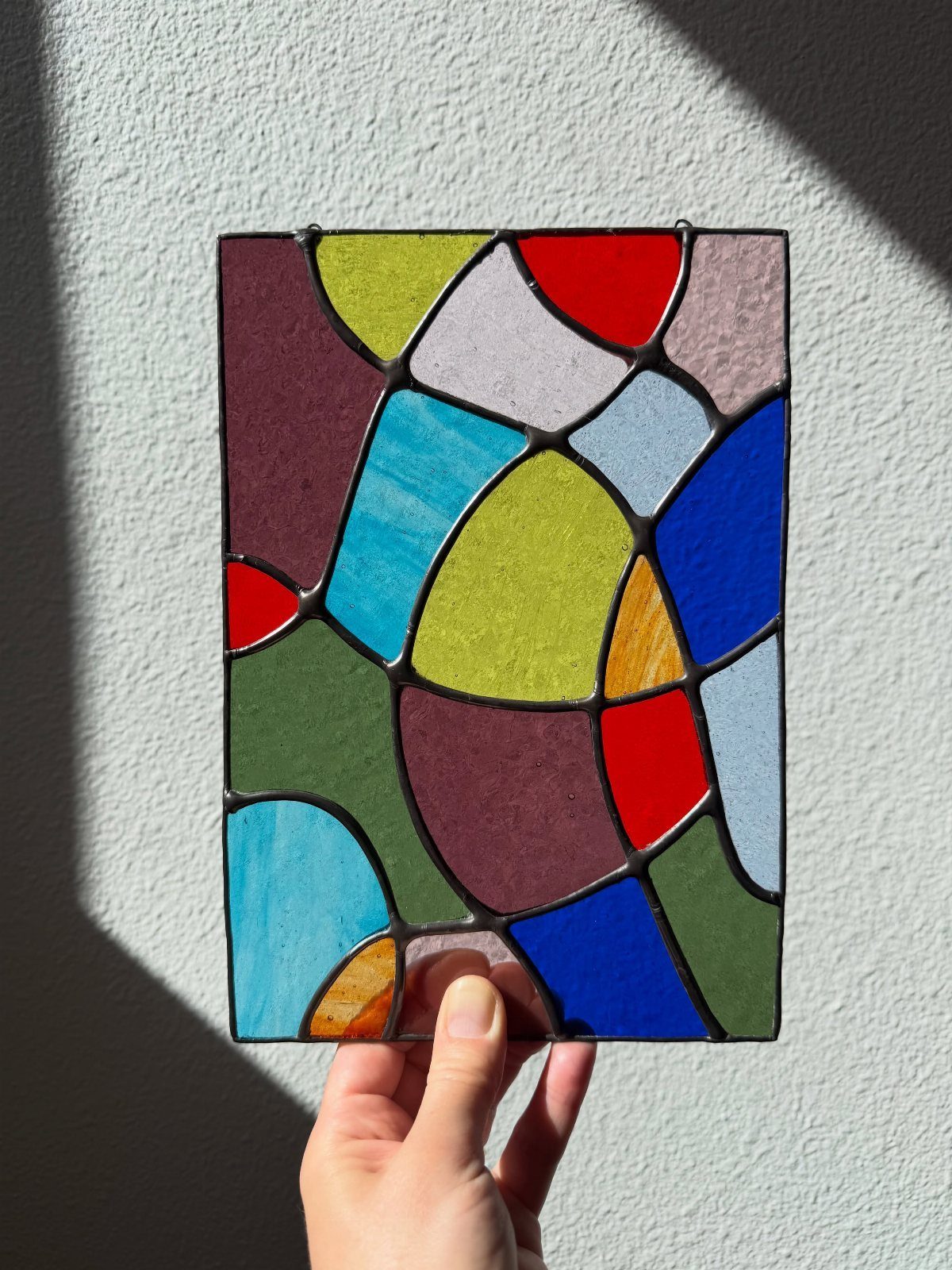 Colorful Stained Glass Design: 10 Ideas to Light a Room 7 A hand holding a rectangular stained glass panel with colorful organic shapes against a white wall.