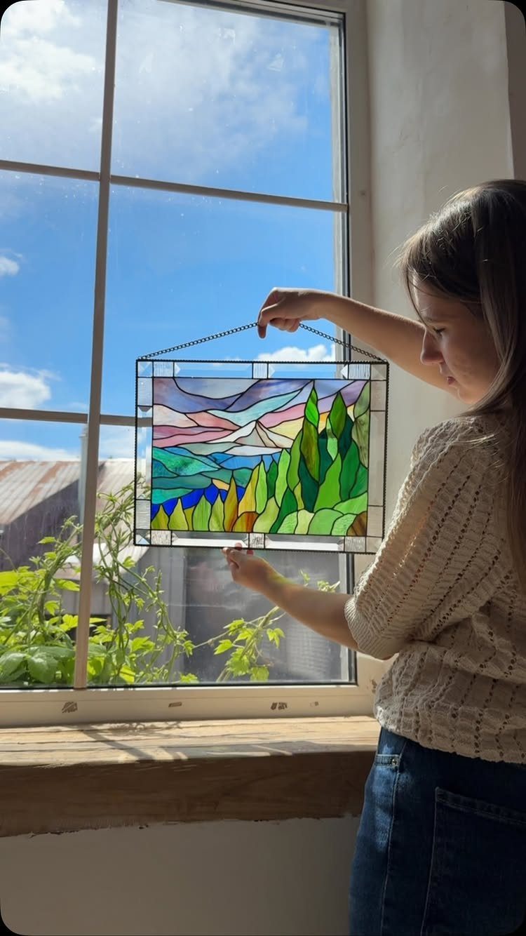 Colorful Stained Glass Design: 10 Ideas to Light a Room 3 A person holding a colorful stained glass landscape featuring green trees and blue mountains against a window.