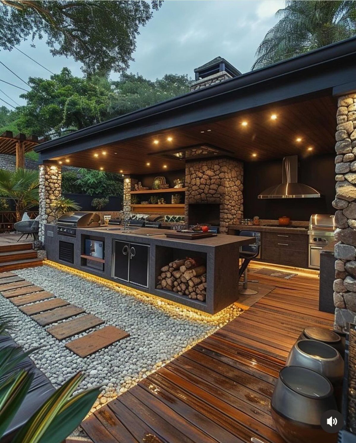 A luxury covered outdoor kitchen with a stone fireplace, dark cabinetry, and a wooden deck.