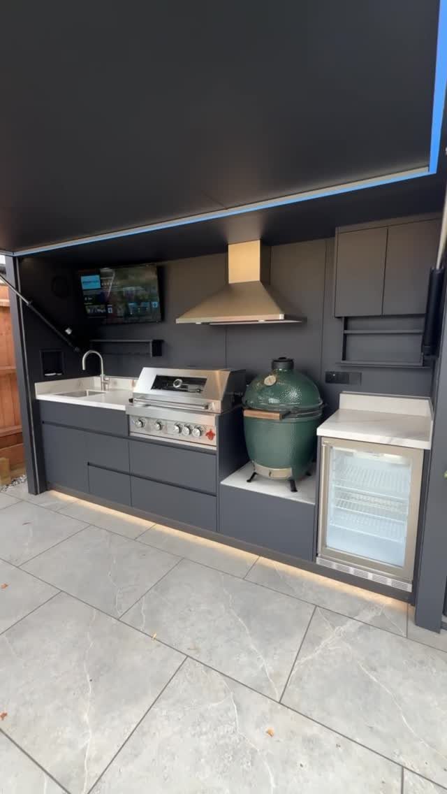 Modern outdoor kitchen with grill, ceramic cooker, sink, and refrigerator under a dark shelter.