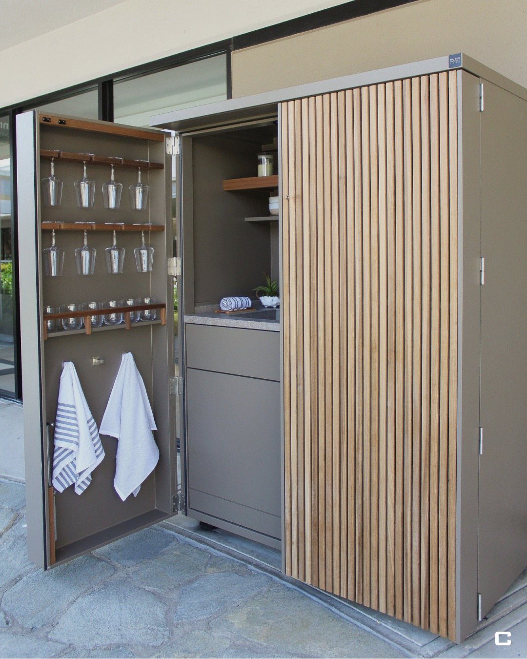 An open outdoor storage cabinet featuring wooden slats, glassware racks, and hanging towels.