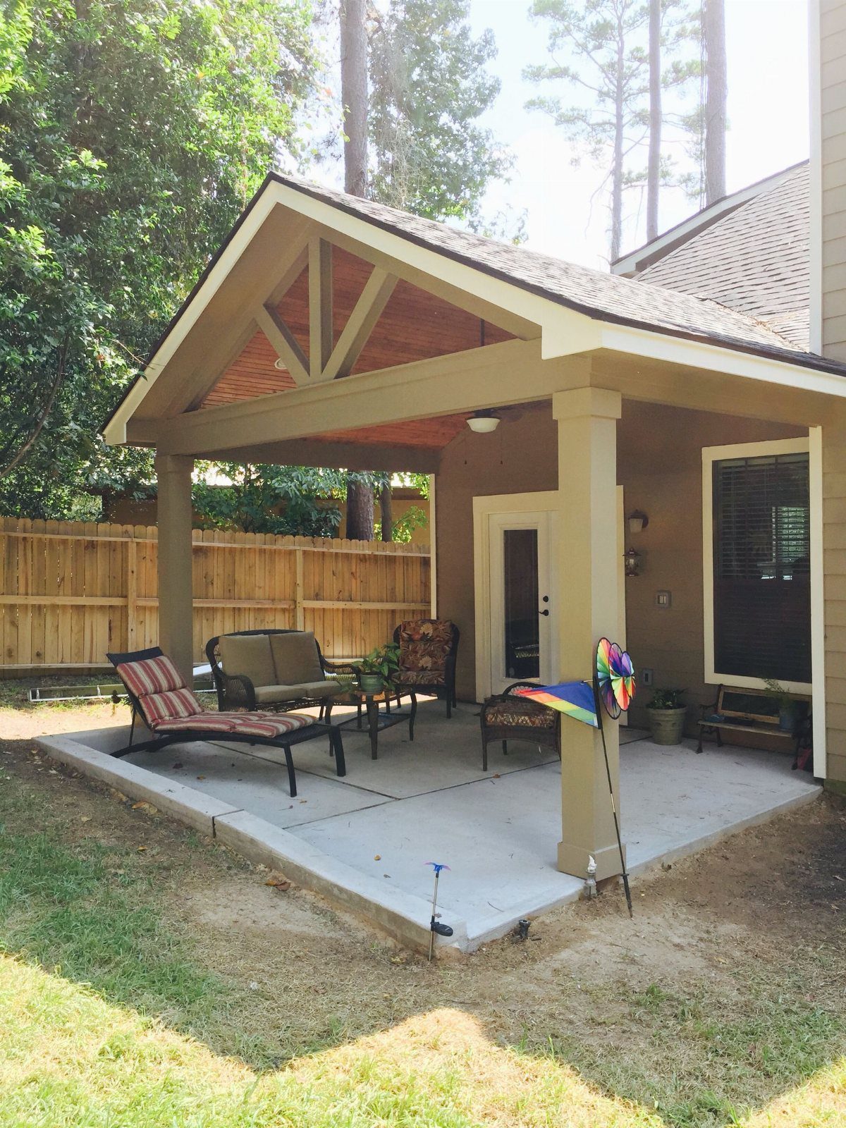 A tan house with a large gabled roof patio cover and outdoor lounge furniture on a concrete patio.