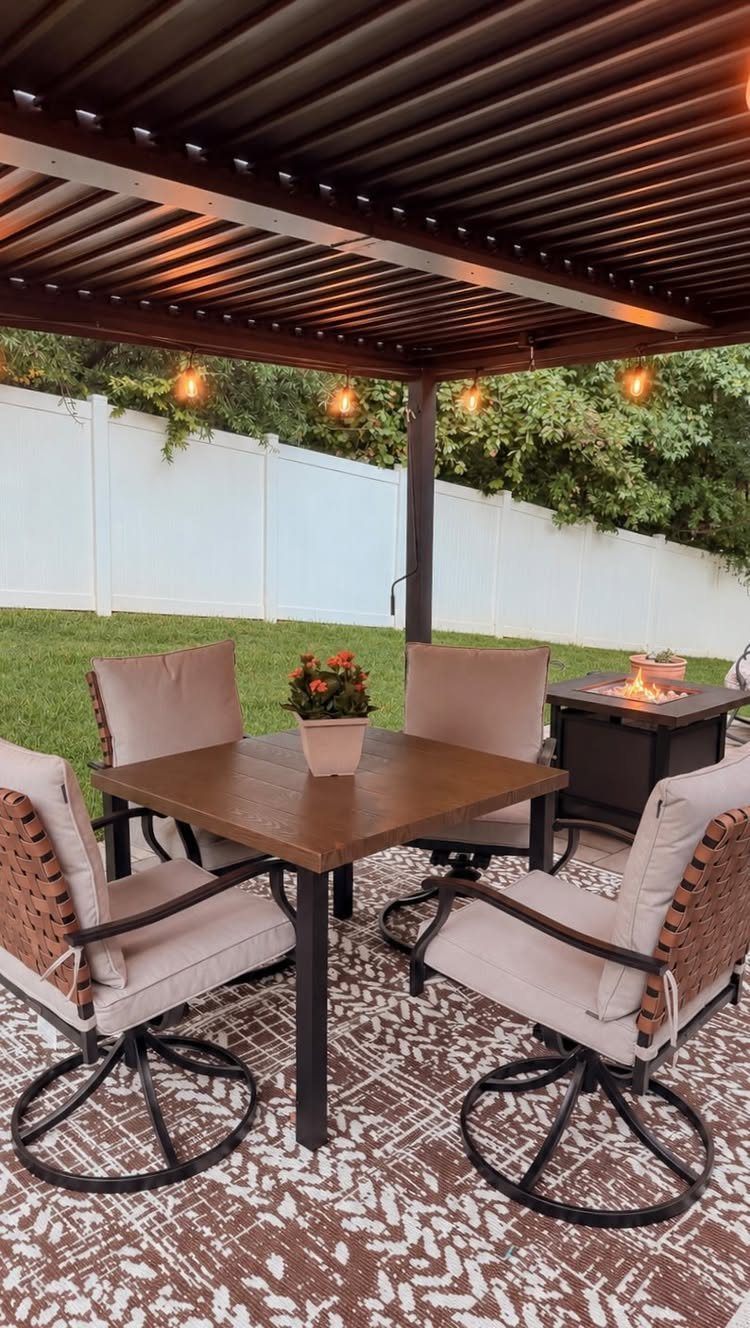 Wooden dining table with four cushioned chairs set on a patterned rug under a louvered patio cover.