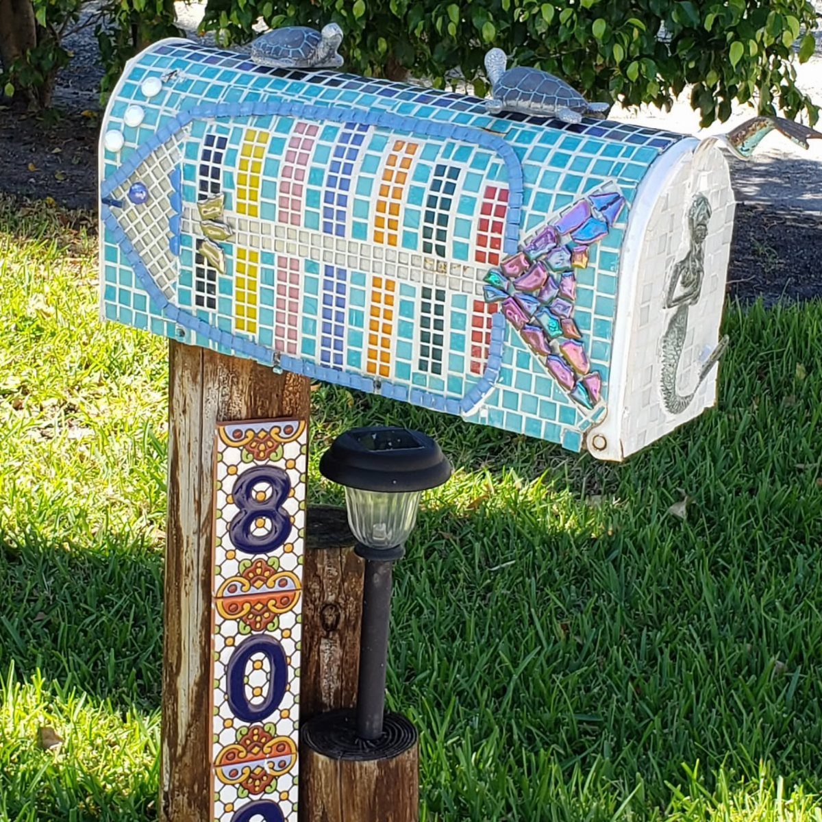 A colorful mosaic-tiled mailbox with turtle figurines sits on a wooden post in a grassy yard.