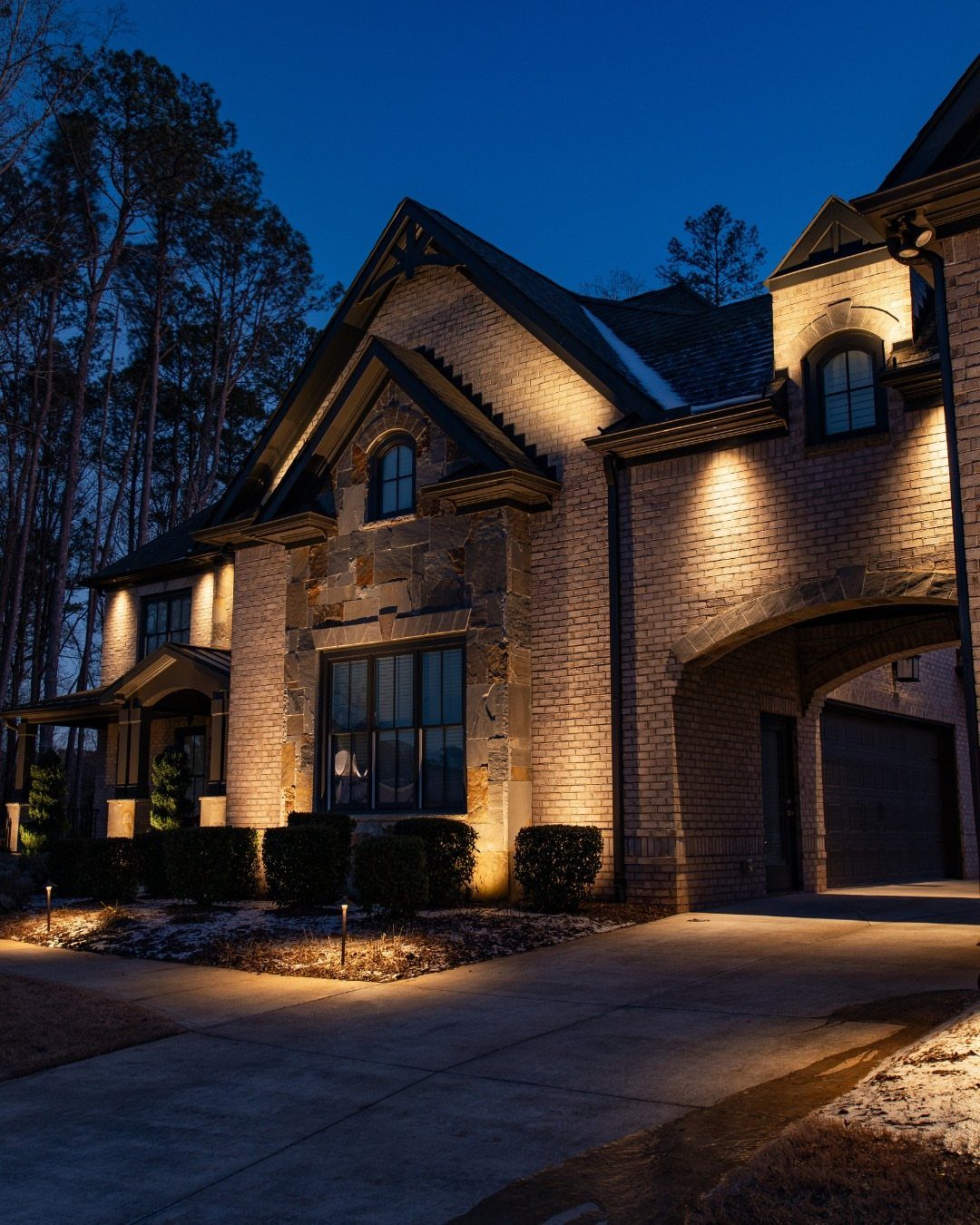 A large brick house illuminated by warm exterior lights along the driveway and facade at dusk.