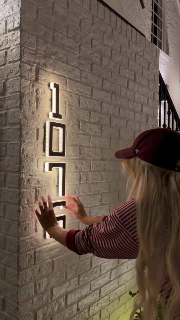 Large illuminated modern house numbers mounted vertically on a white brick wall at night.