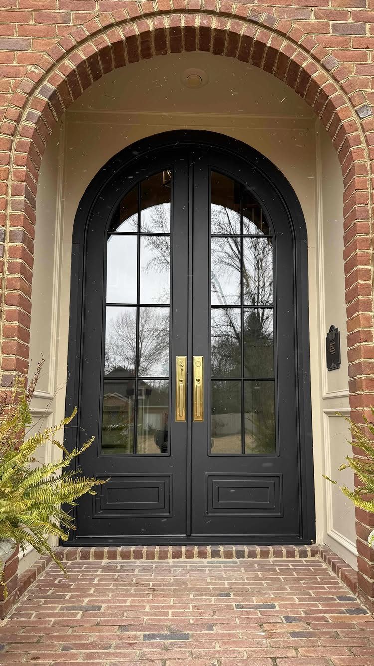 Black arched double doors with glass panes and gold handles set in a brick entryway.