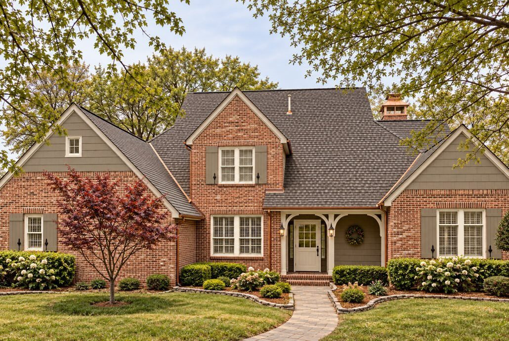 A brick house with olive green shutters and matching window frames surrounded by green landscaping.