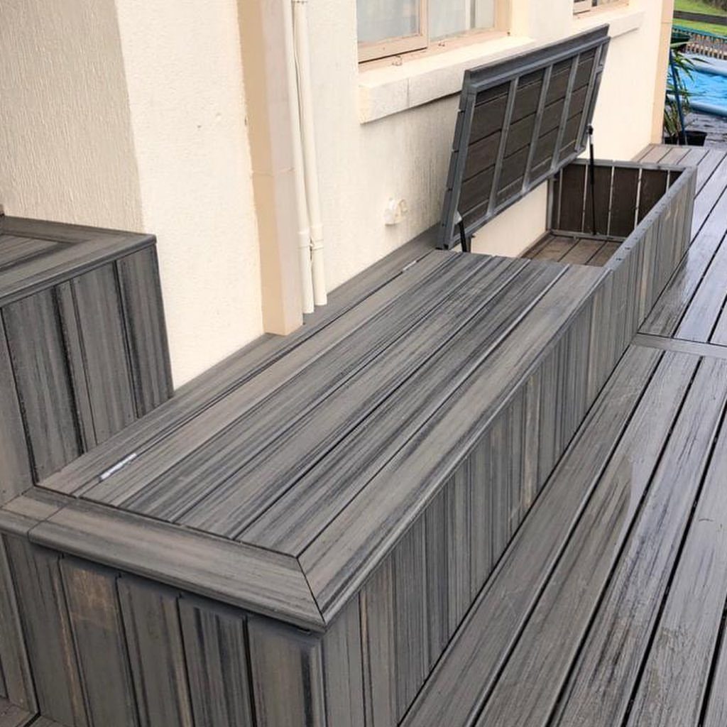 Grey wooden deck with an open built-in storage bench compartment near a building wall.