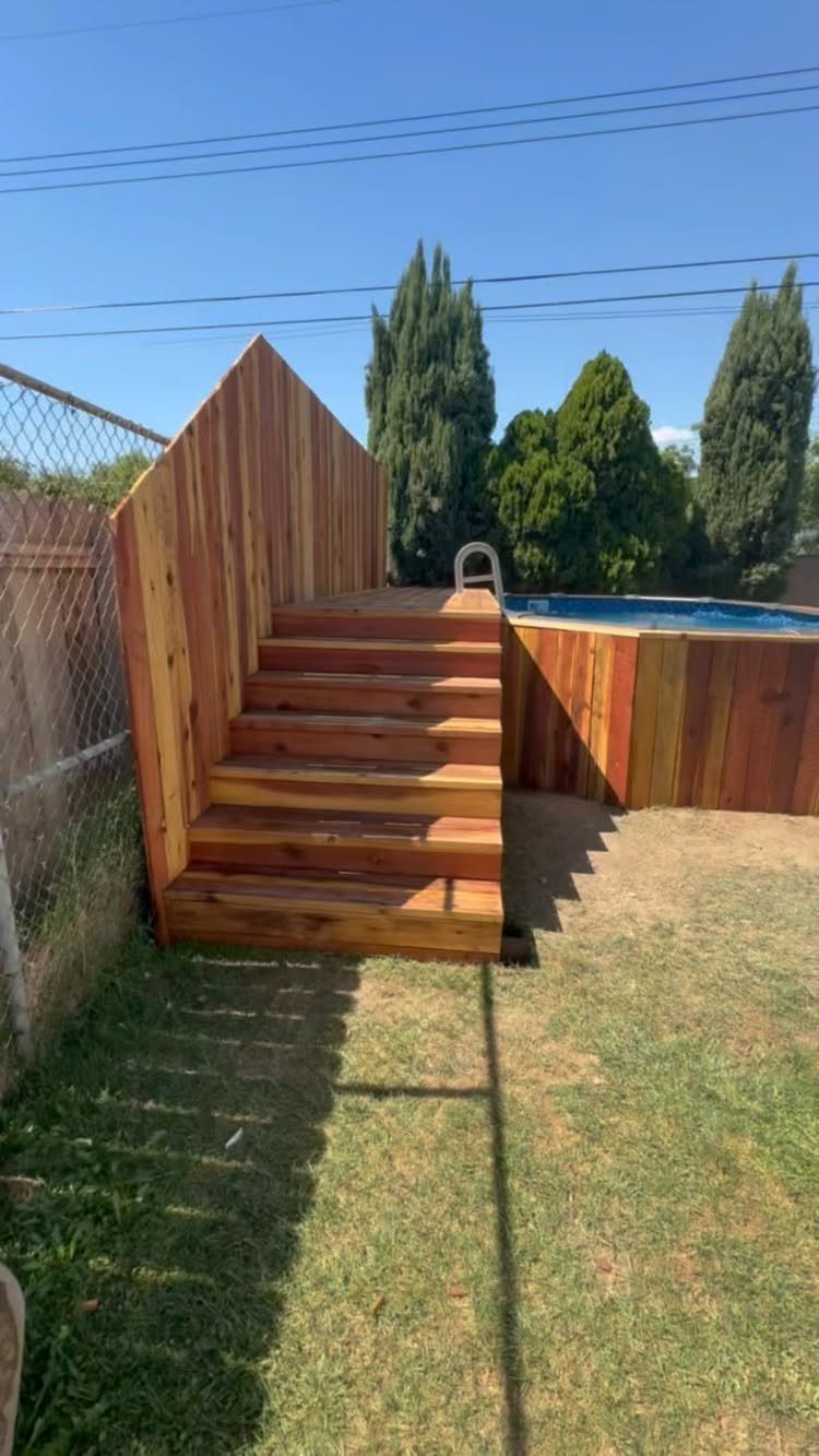 Wooden deck with stairs surrounding an above-ground pool in a grassy backyard.