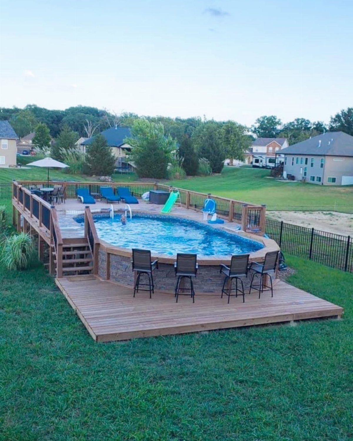 Wooden deck surrounding an above-ground pool with blue lounge chairs and bar stools on a lawn.