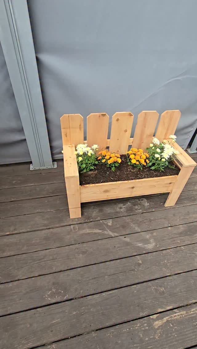 A light wood planter box with a picket fence backrest containing orange and white flowers.