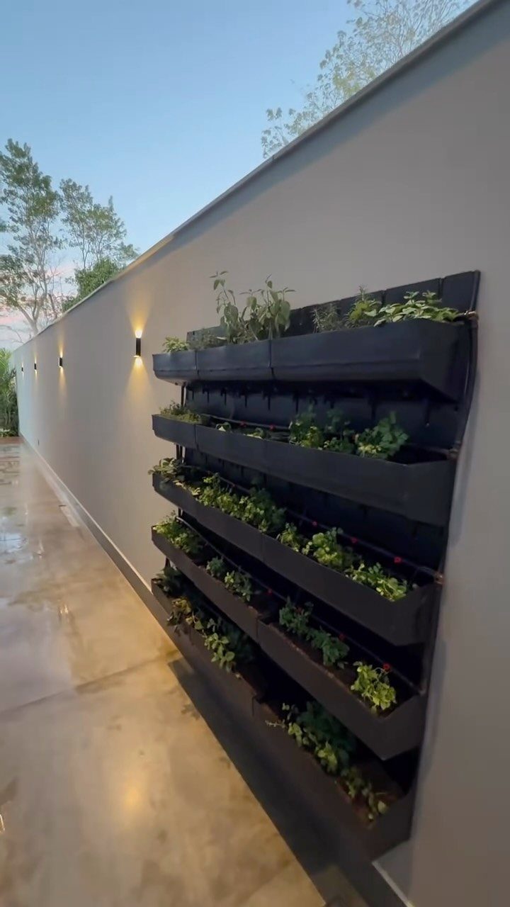 Black tiered vertical garden planter with green plants mounted on a grey outdoor wall.