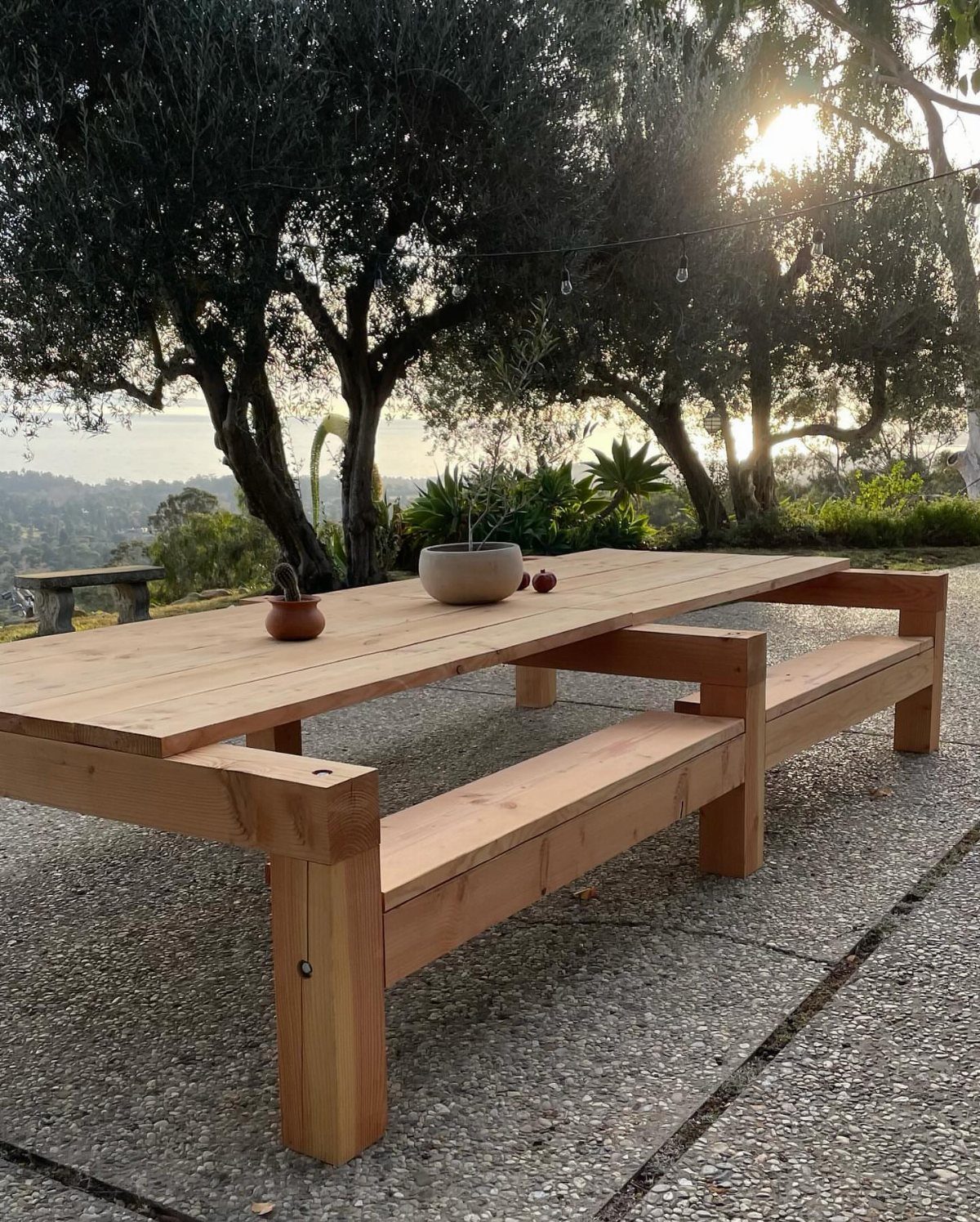 Easy Patio Table DIY: 8 Projects for Your Yard 4 A long wooden outdoor dining table with integrated benches on a stone patio under trees.