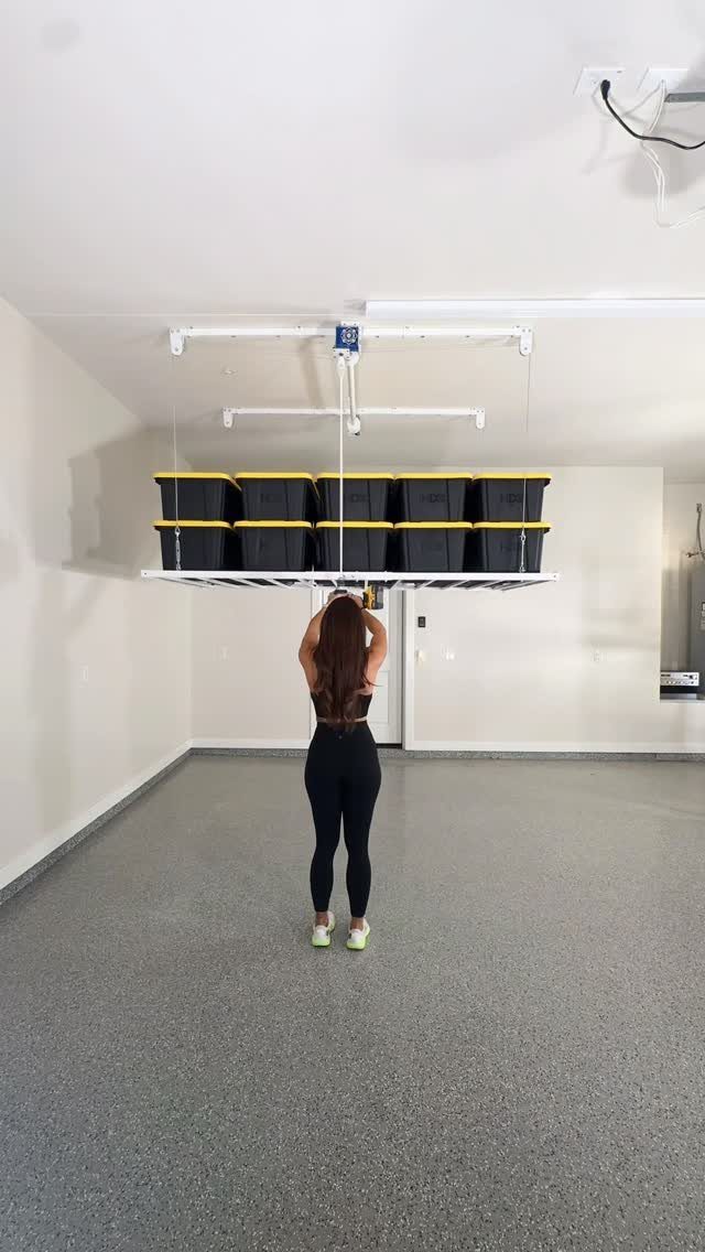 A woman stands in a garage looking up at a ceiling-mounted storage rack with black bins.