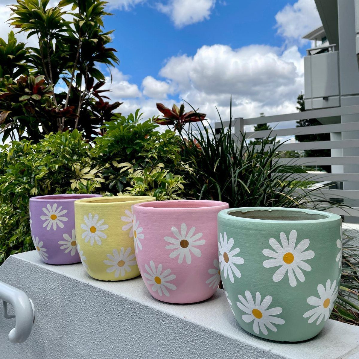 Four pastel-colored ceramic pots with white daisy patterns sitting on a white ledge outdoors.