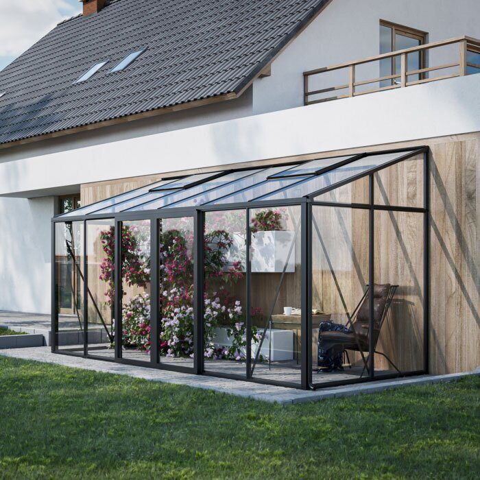A modern black-framed glass greenhouse attached to a house, containing pink flowers and a small seating area.