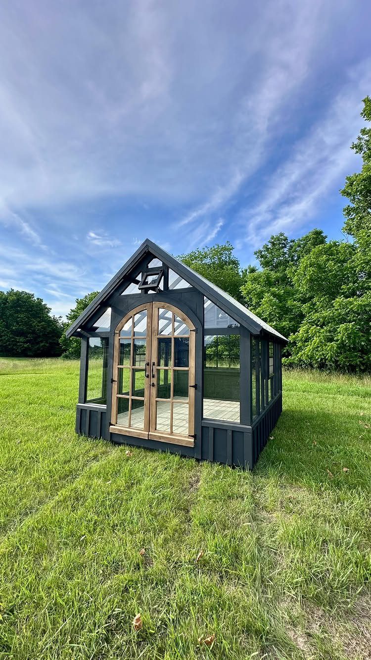 A black-framed glass greenhouse with wooden arched doors sits in a lush green field under a blue sky.