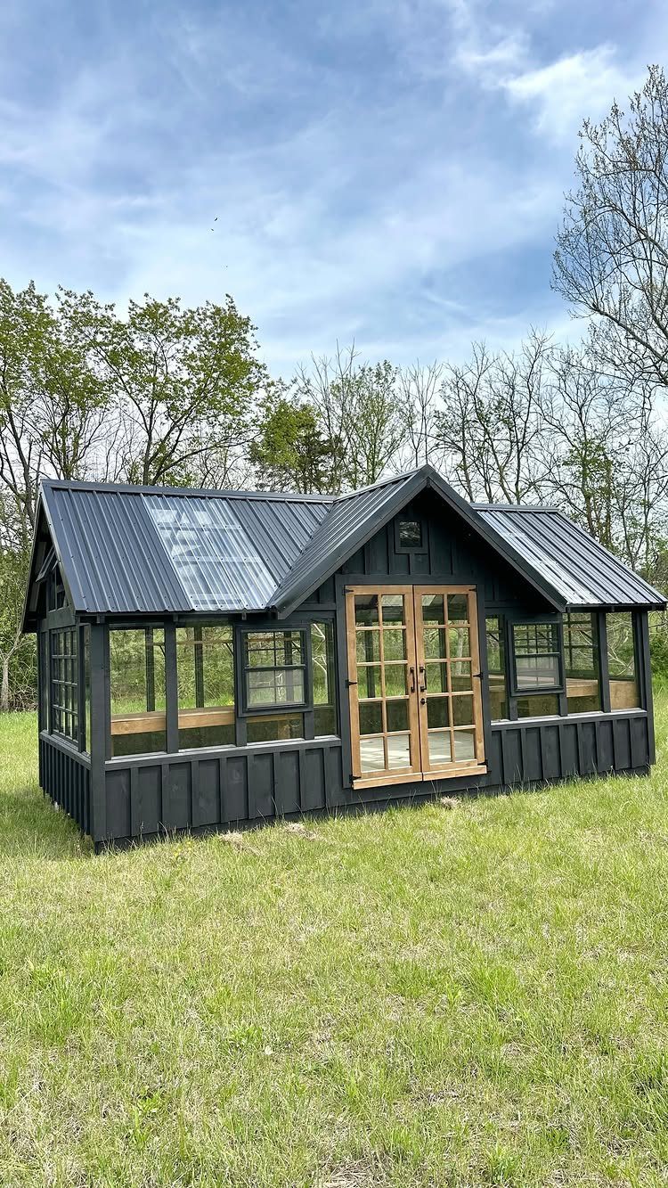Black wooden gable greenhouse with large glass windows and doors on a green lawn.
