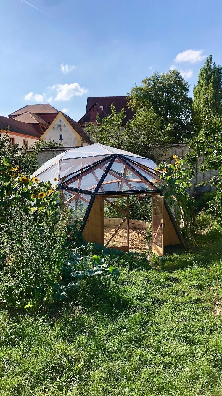 A geodesic dome greenhouse with white sheeting and a wooden door in a lush garden.
