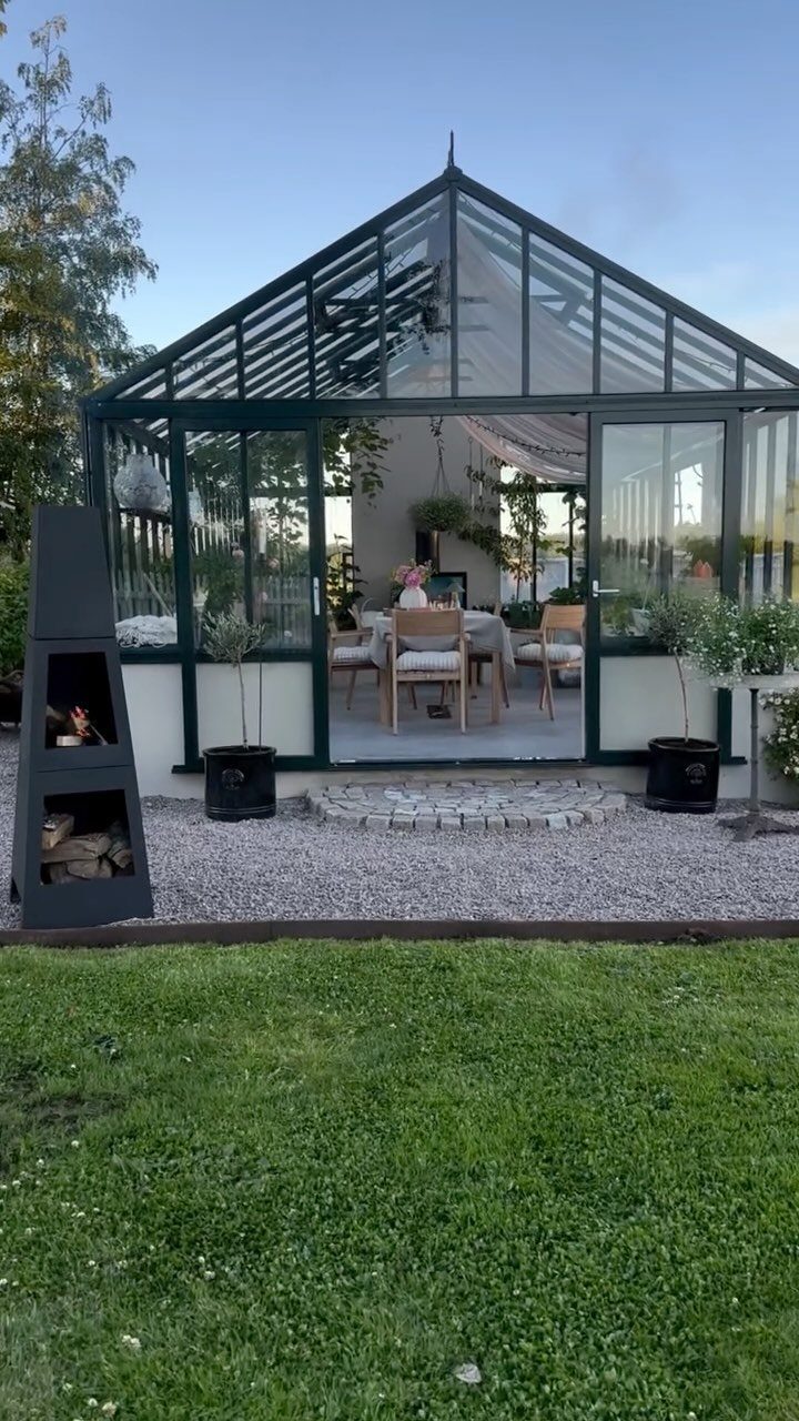 A modern dark green glass greenhouse containing a dining set on a gravel patio.
