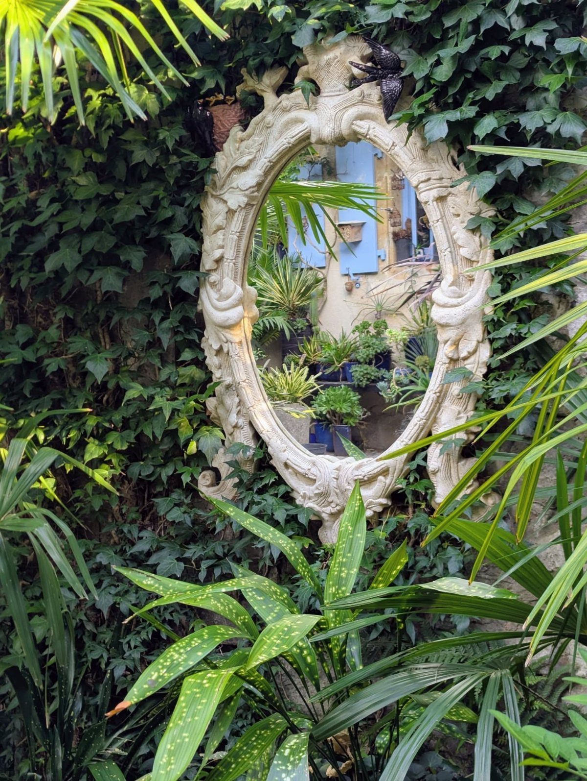 21 Home Decor Ideas for Garden Spaces That Work in Any Yard 18 Ornate white mirror reflecting a sunny garden with plants and blue shutters amidst green ivy.