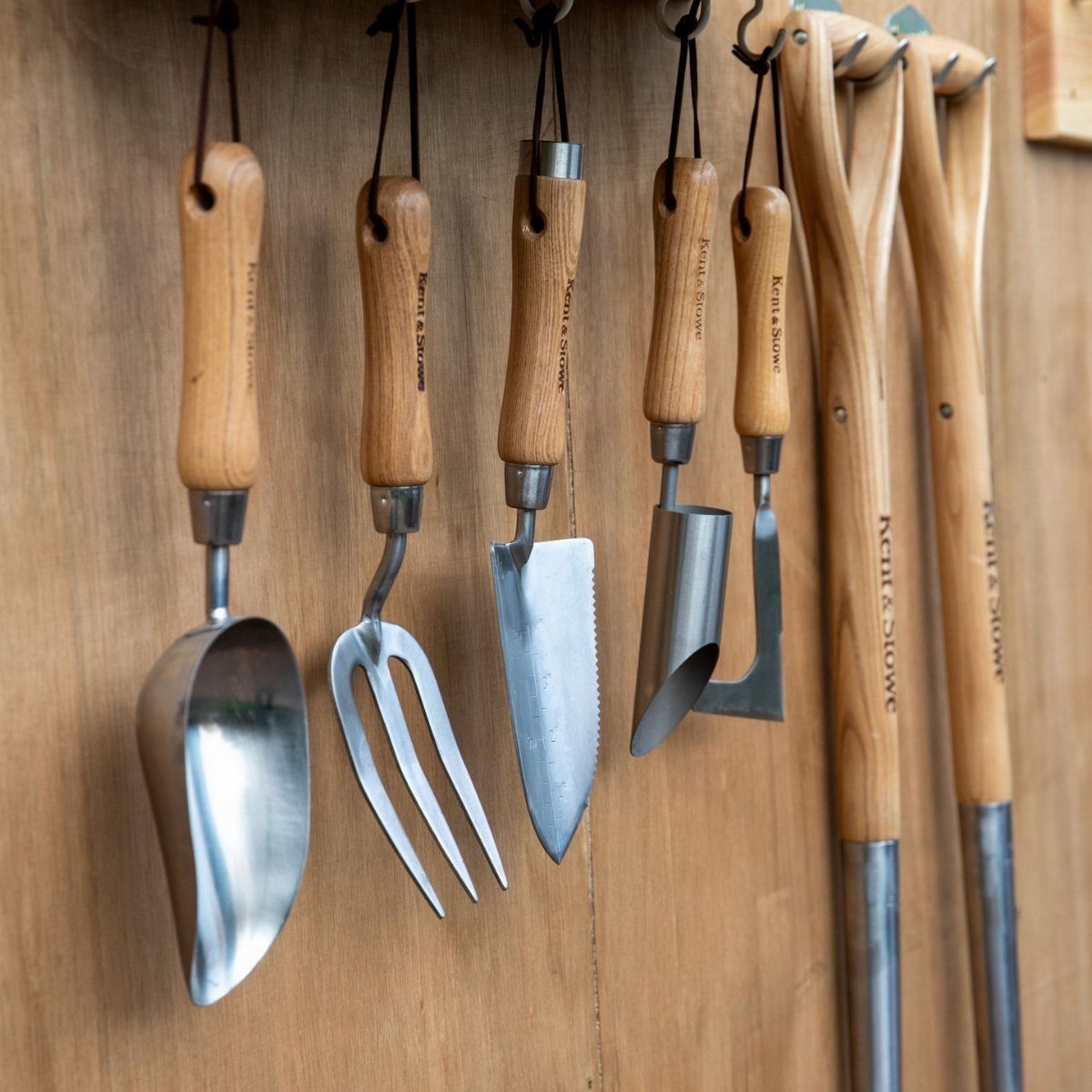 Various metal hand gardening tools with wooden handles hanging against a wooden wall.